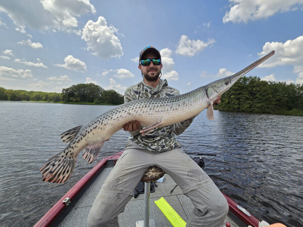 Un homme assis sur un bateau de pêche rouge sur un lac tenant un lépisosté, un long poisson gris tacheté et beige.