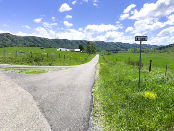 Un paysage rural pittoresque avec une route pavée qui bifurque dans deux directions. Sur la gauche, la route débouche sur un champ verdoyant avec des collines en arrière-plan, tandis que sur la droite, la route continue tout droit vers une ferme lointaine. Un panneau indiquant "608" est visible sur le côté droit de la route. Le ciel est d'un bleu éclatant, parsemé de nuages blancs et cotonneux.