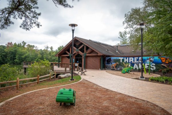 L'entrée principale du Centre Nature, qui est un bâtiment marron sur un côté duquel est peinte une fresque murale qui indique « Trois Lacs ». À gauche du bâtiment se trouve une terrasse d'observation en bois qui donne sur l'un des lacs.